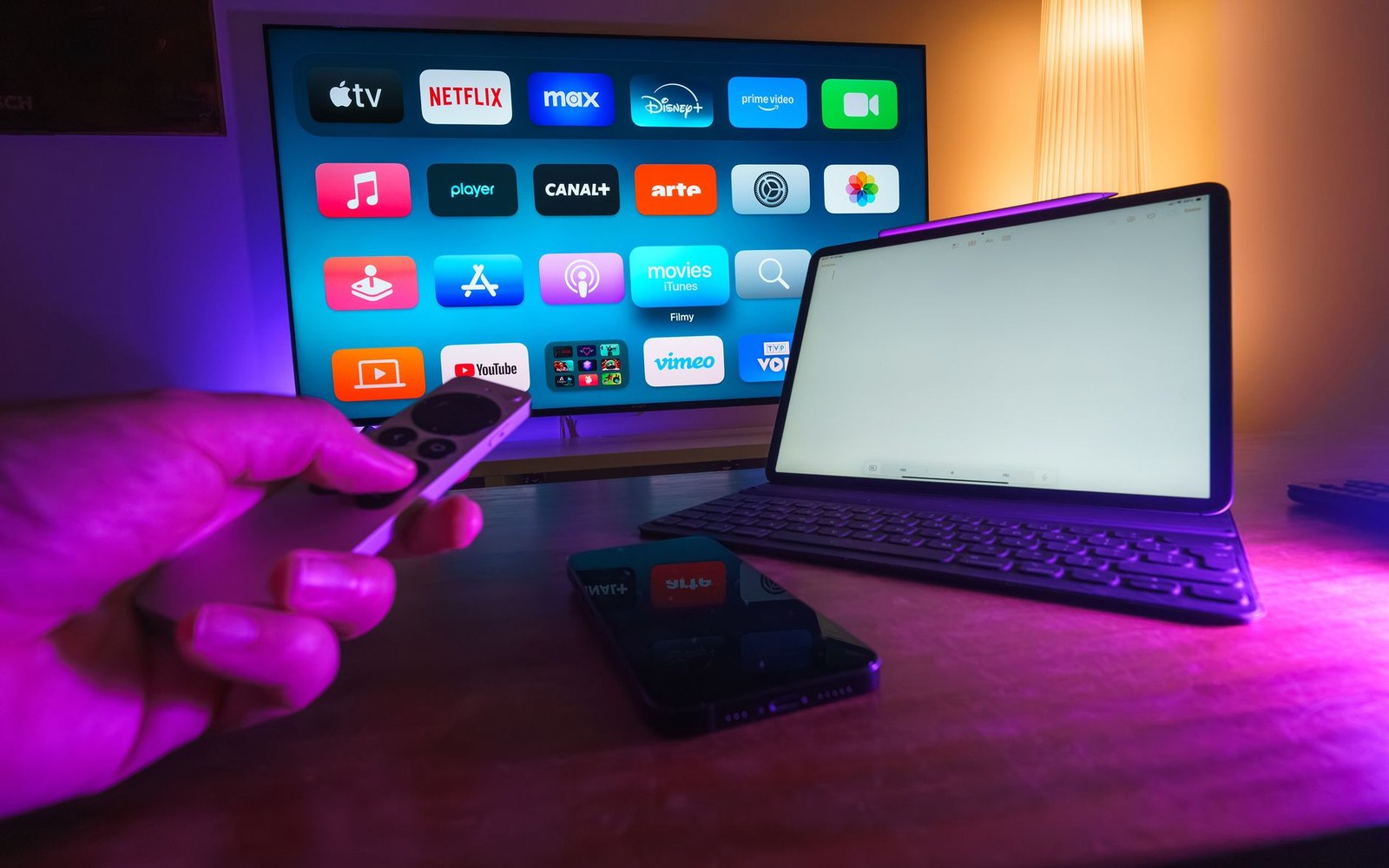 A hand holds a streaming remote toward a smart TV app menu, with a tablet and phone nearby
