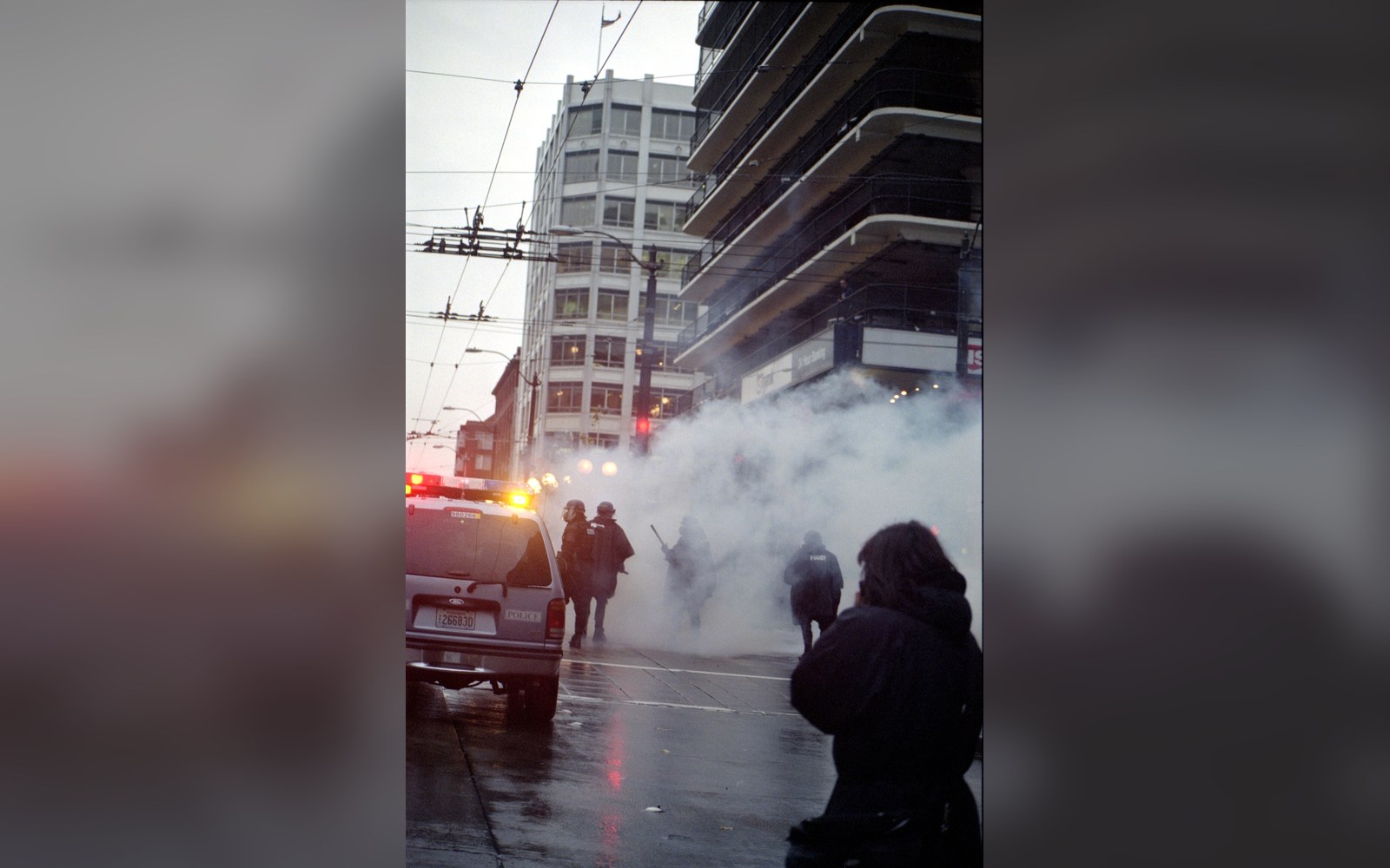 Riot police stand near a patrol car amid tear gas smoke on a rainy street