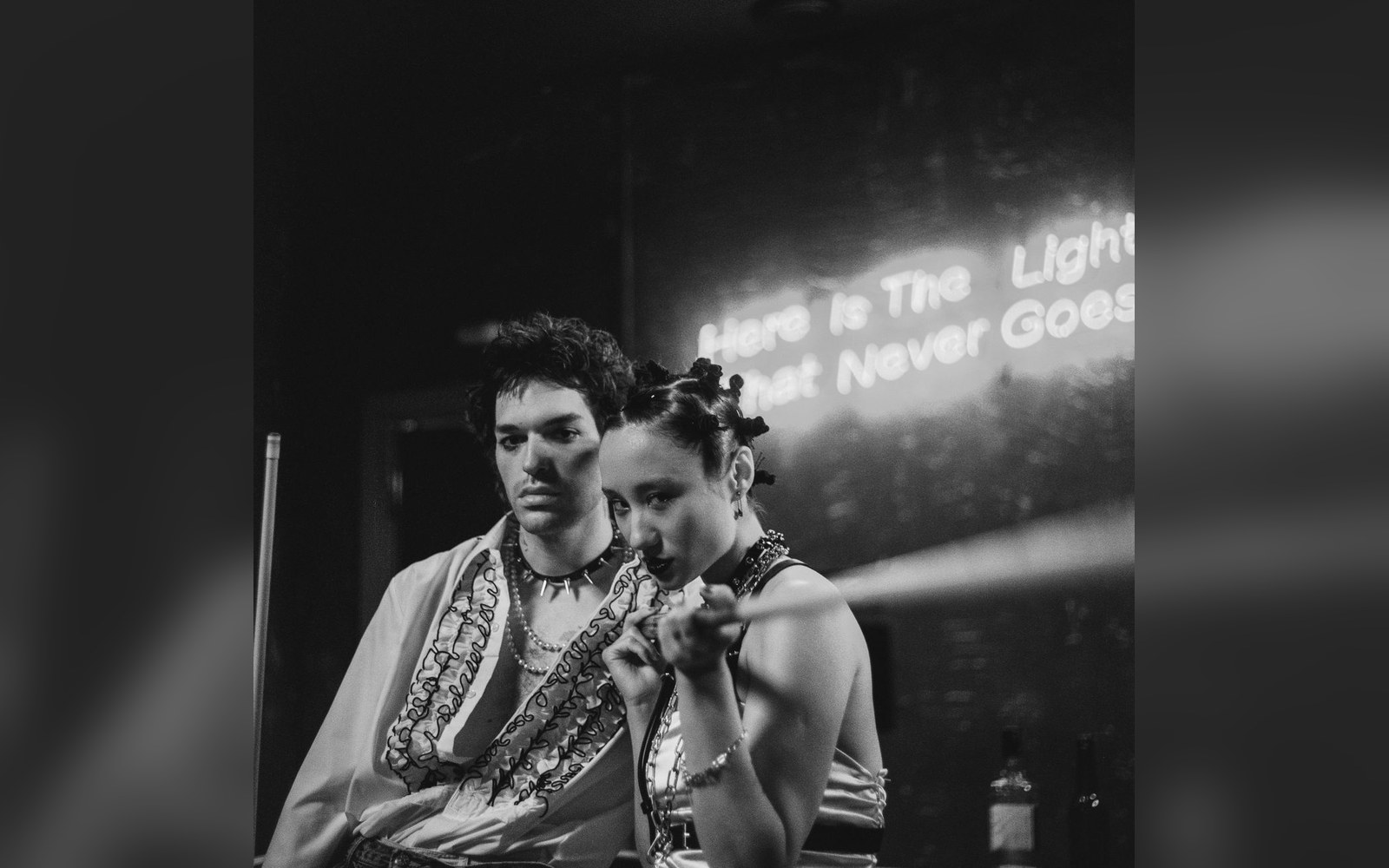 Man and woman posing near a neon sign reading "here is the light that never goes"