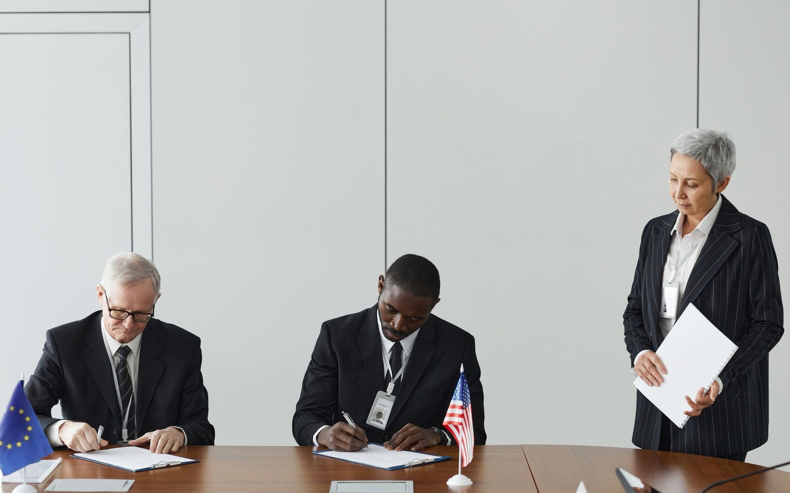 Two officials sign documents at a table with EU and US flags as a woman looks on