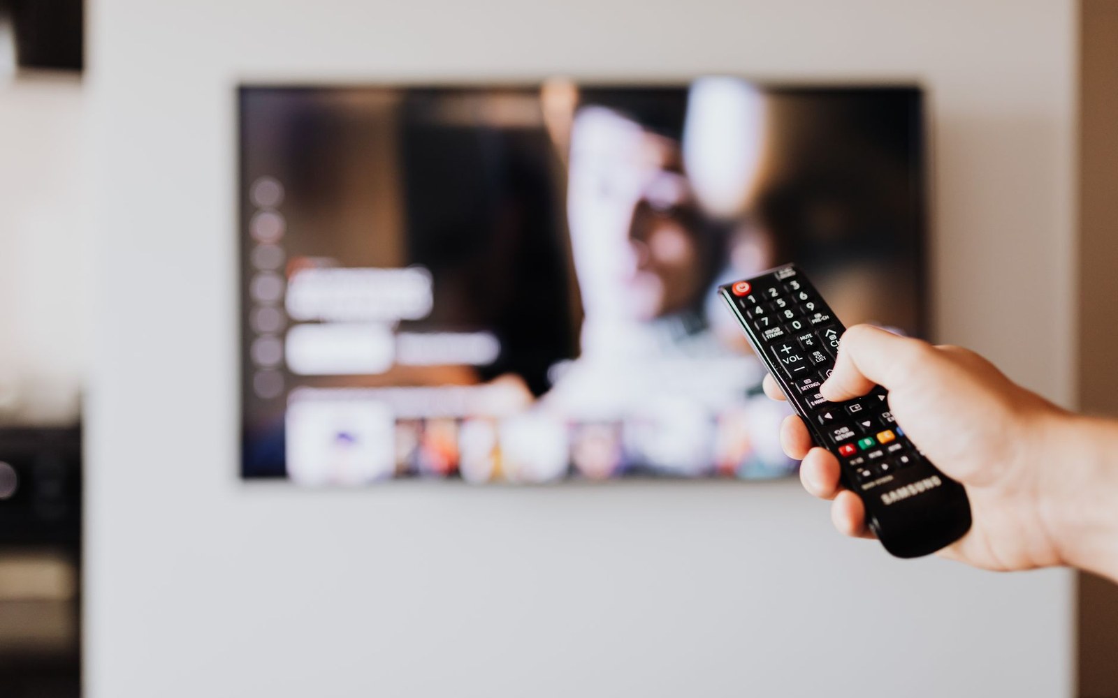 A hand points a Samsung remote control at a wall-mounted television