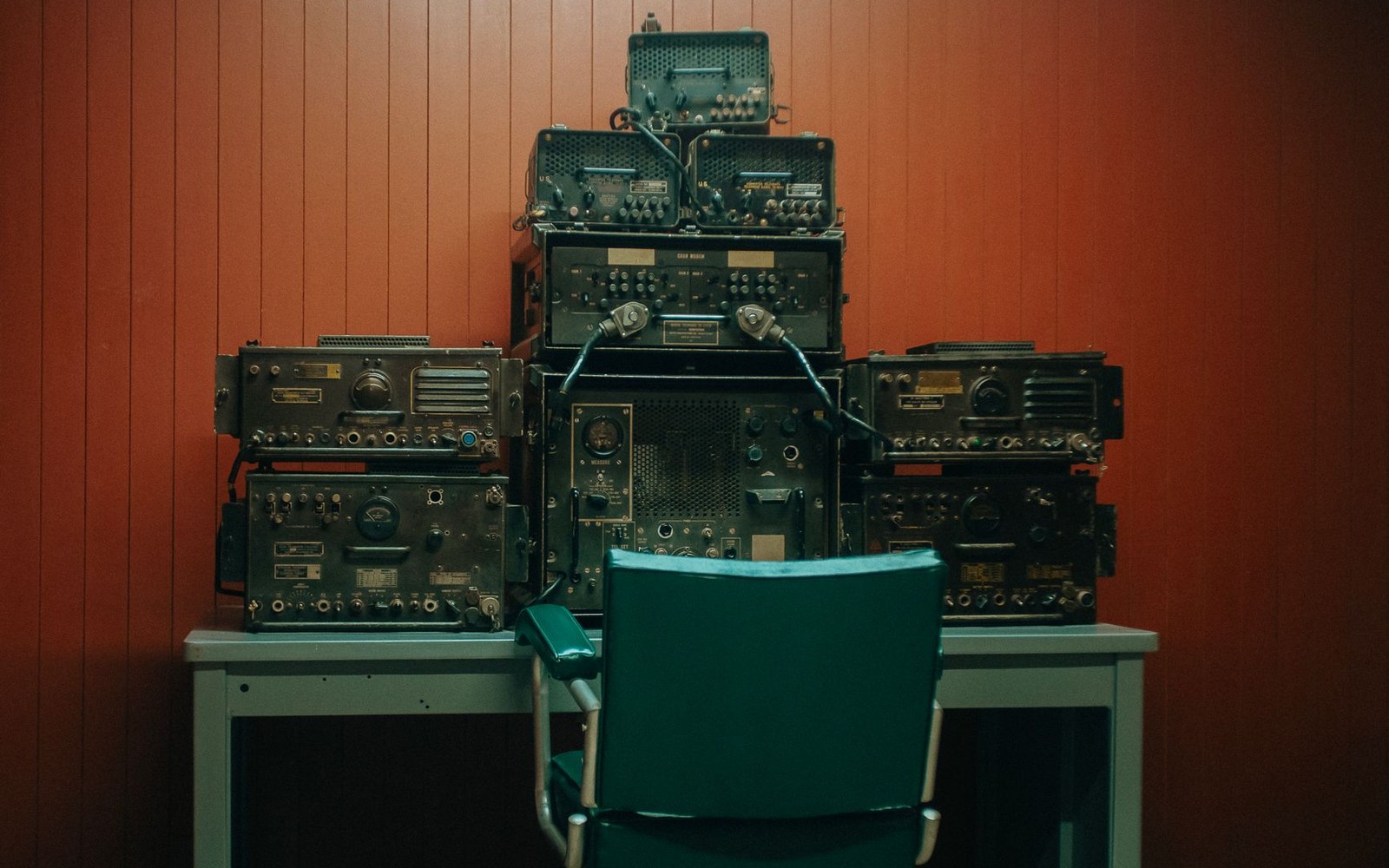 Stacked vintage military radio equipment on a desk with an empty teal chair