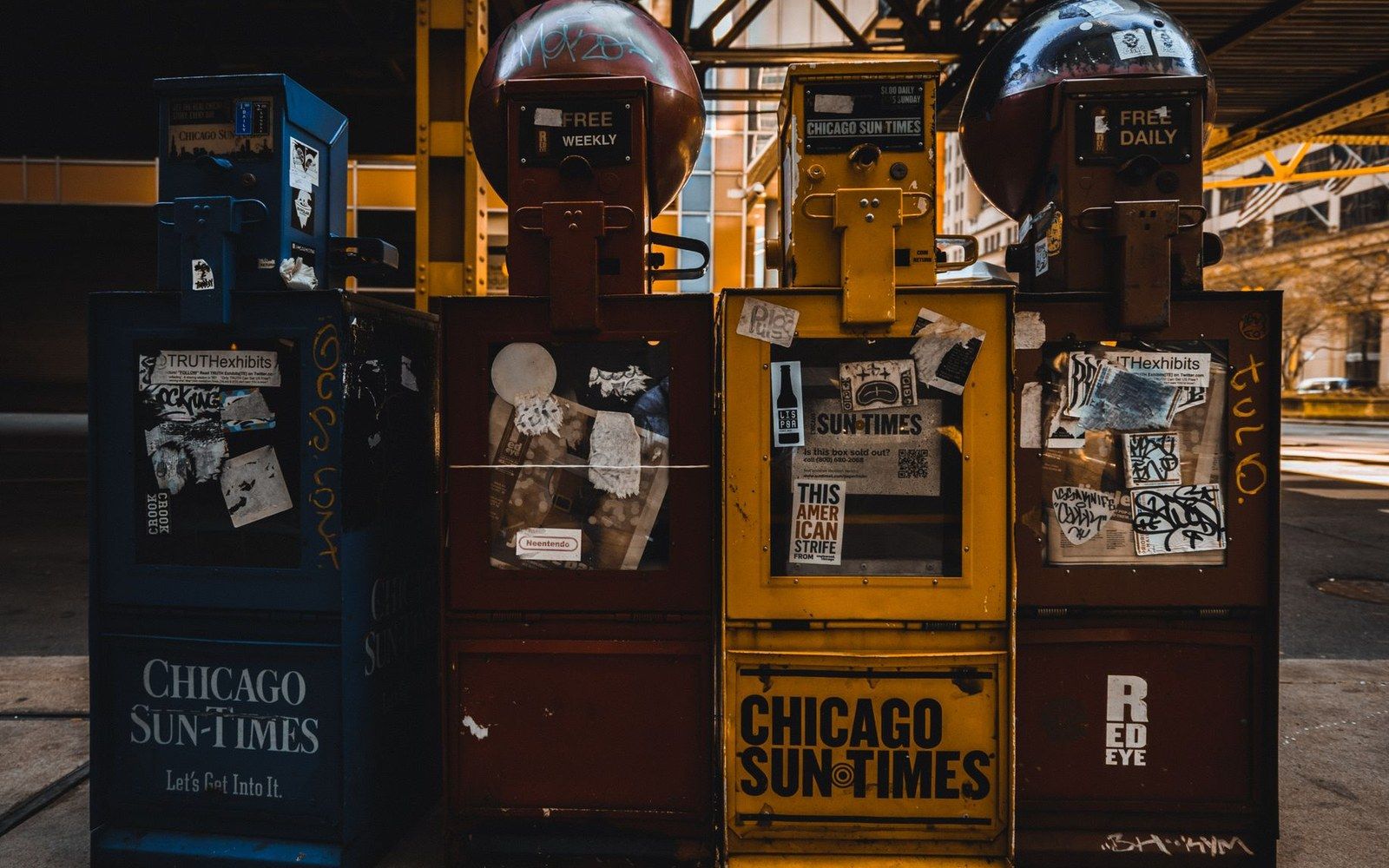 Row of weathered Chicago Sun-Times and Red Eye newspaper vending boxes covered in stickers