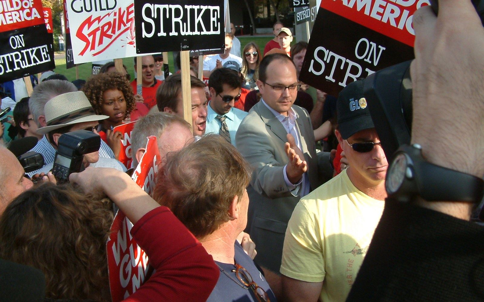 A crowd holds "on strike" and "guild" picket signs while cameras capture the scene
