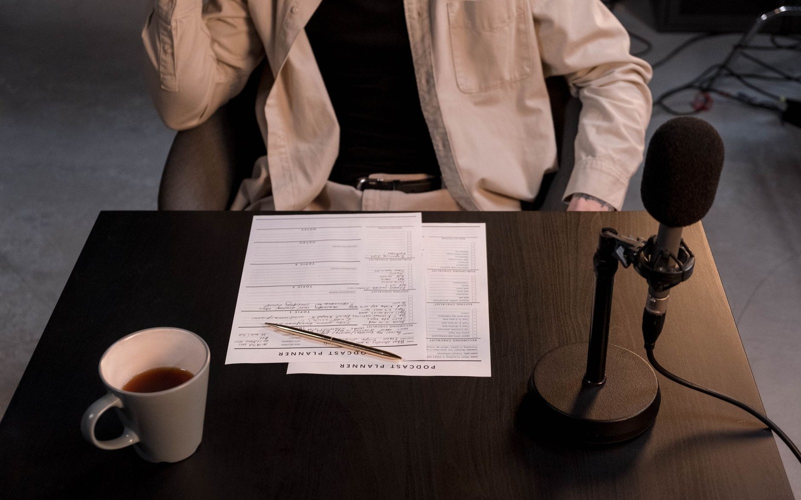 An adult host seated at a home studio desk with a large-diaphragm microphone in foreground